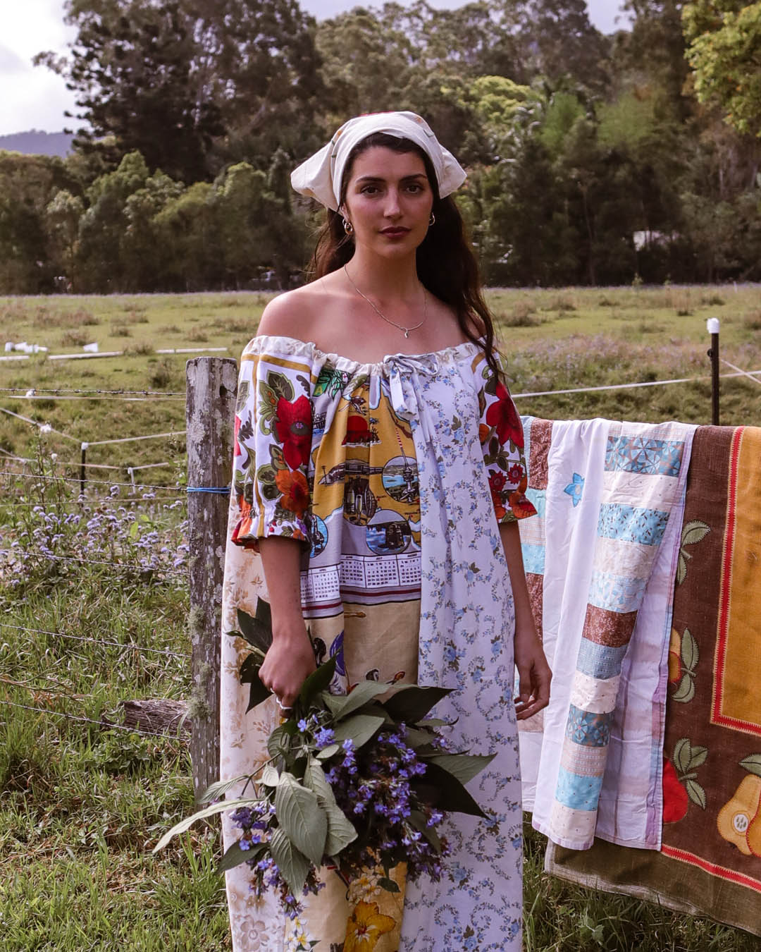 A woman stands outdoors in a grassy field wearing a colorful off-the-shoulder patchwork dress with floral and vintage-style prints, a white head wrap, and holding a bouquet of purple flowers. A quilted blanket is hanging on a fence behind her.