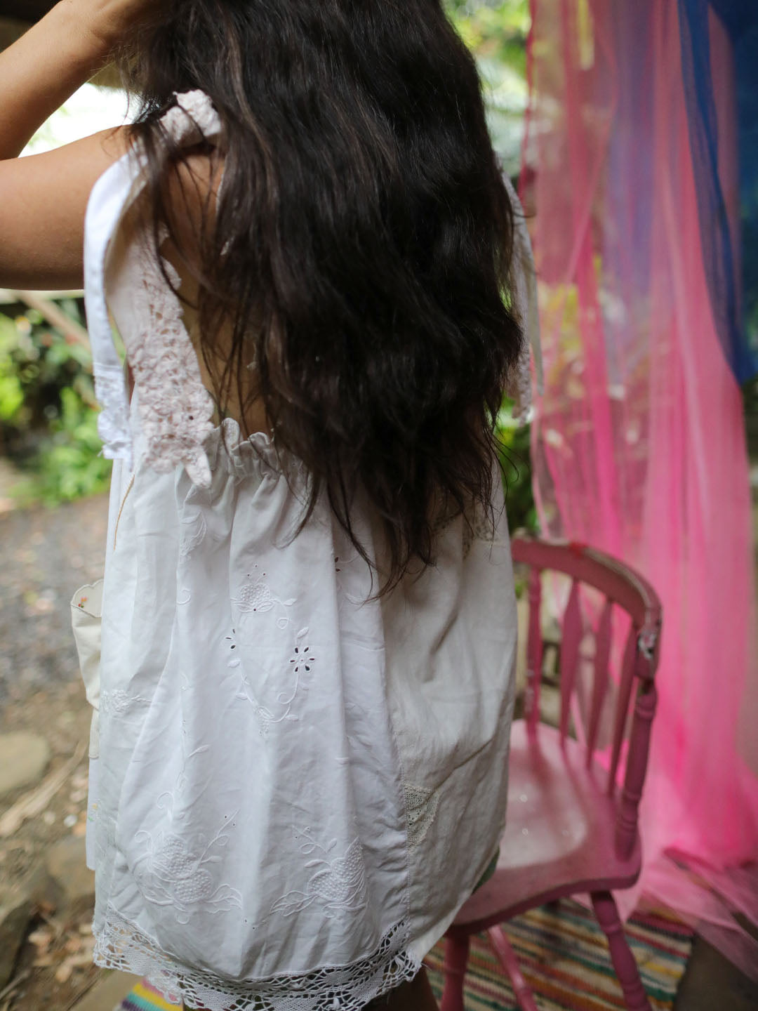A woman viewed from behind wearing a white eyelet sleeveless top, standing next to a pink chair with colorful sheer fabric in the background.
