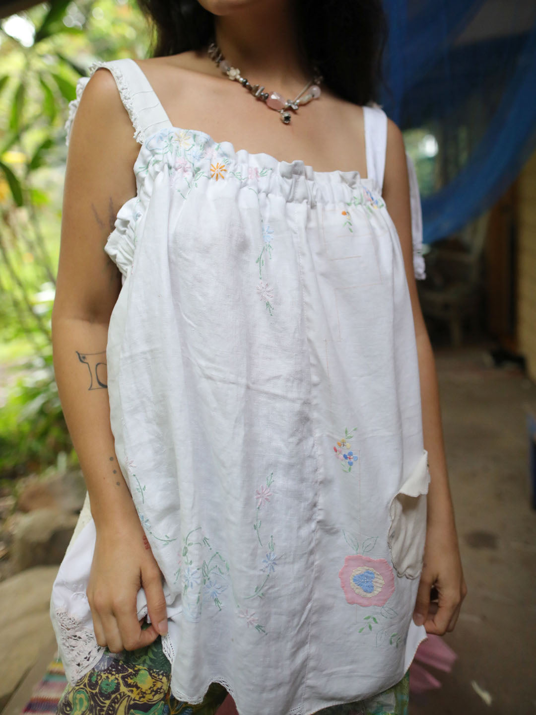 A woman wearing a white cotton camisole top with delicate pastel floral embroidery and ruffled trim, photographed in an outdoor setting with green foliage in the background.