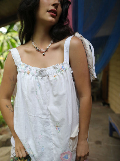 A woman wearing a white sleeveless top with delicate pastel floral embroidery and ruffled trim, photographed in an outdoor setting with a blue curtain in the background.
