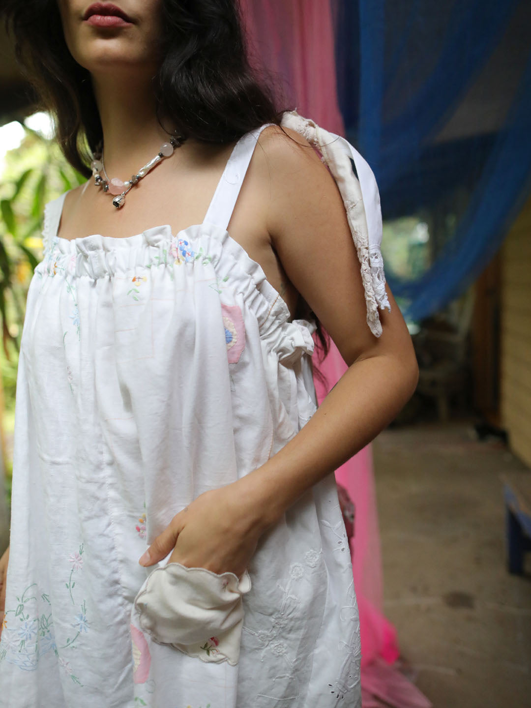 A woman wearing a white sleeveless dress with floral embroidery, ruffled trim, and tie details on the straps, photographed in an outdoor setting with colorful fabric drapes in the background.
