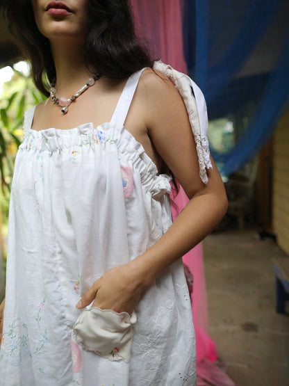 A woman wearing a white sleeveless dress with floral embroidery, ruffled trim, and tie details on the straps, photographed in an outdoor setting with colorful fabric drapes in the background.