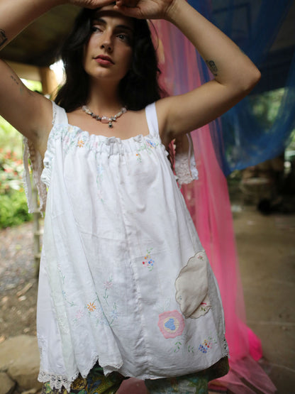 A woman wearing a white sleeveless embroidered top with lace trim and a patch pocket, standing in an outdoor rustic setting with a pink fabric backdrop.