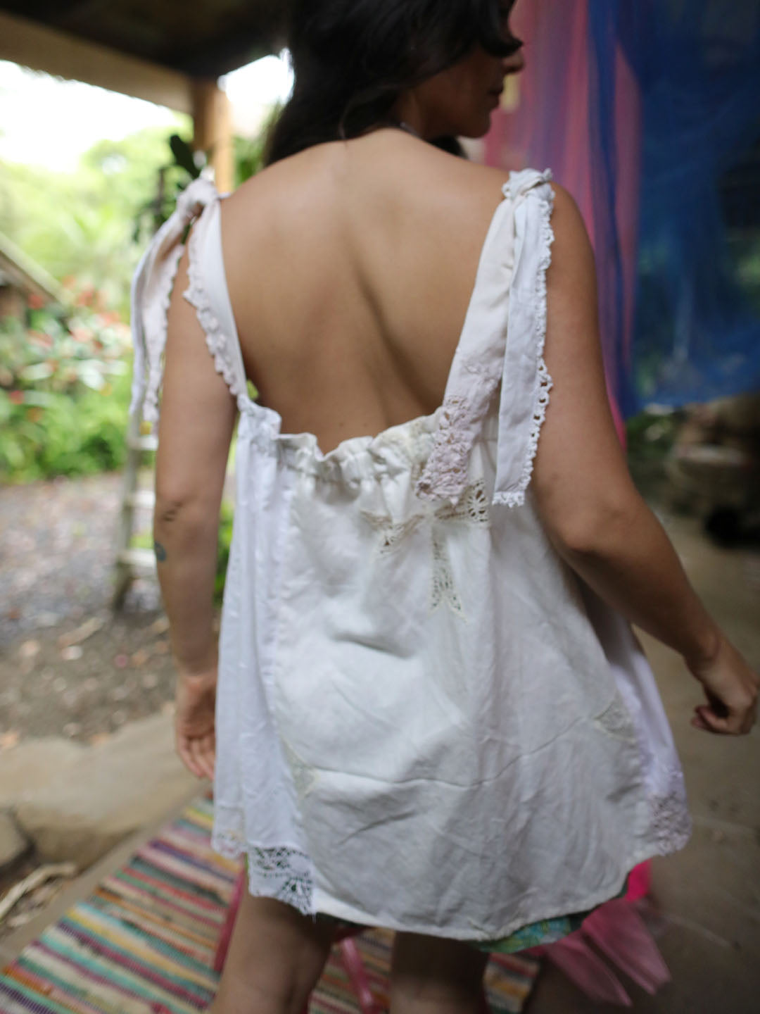 A woman viewed from behind wearing a loose white cotton slip or nightgown with lace trim and tie straps, photographed in an outdoor porch setting.