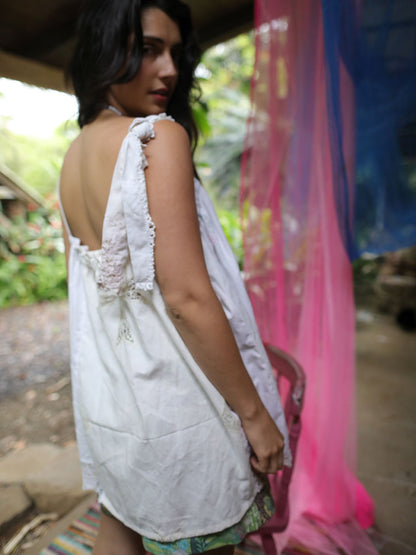 A woman wearing a white sleeveless cotton nightgown or tunic with lace trim, photographed from the back in an outdoor rustic setting with colorful pink and blue fabric hanging nearby.