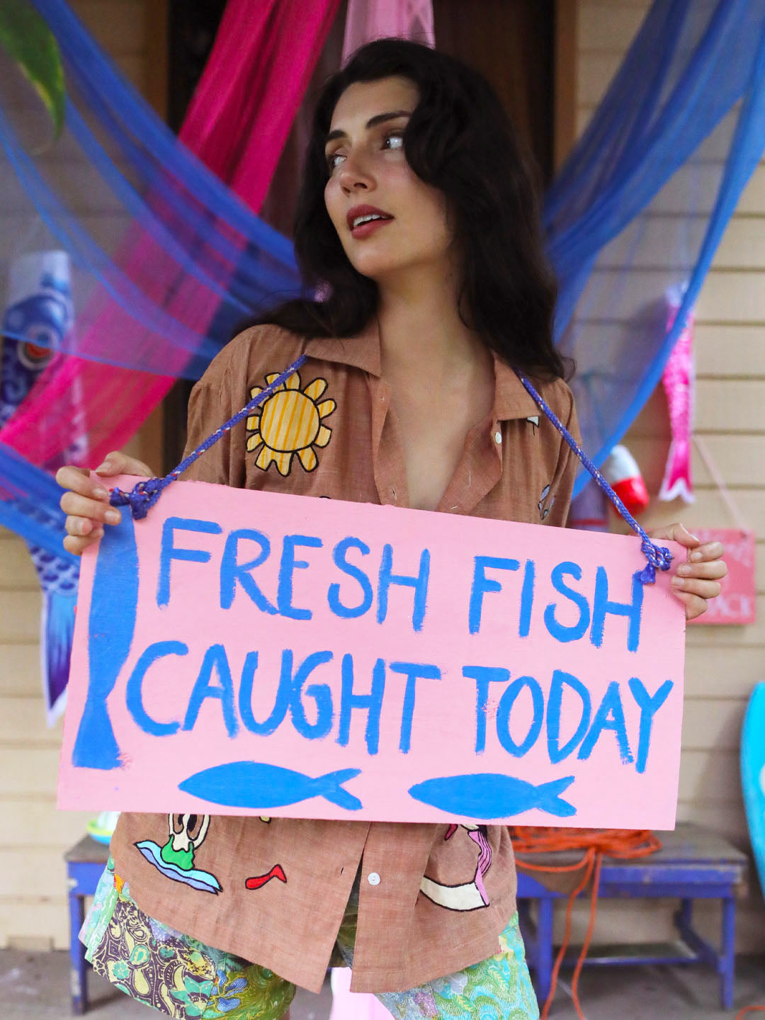 A woman holding a pink hand-painted sign that reads 'FRESH FISH CAUGHT TODAY' in blue letters, with two blue fish illustrations at the bottom. She is wearing a brown shirt with a yellow sun patch and colorful patterned shorts, standing in front of a house with blue hammocks.