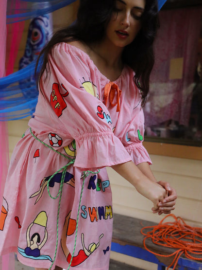 A woman wearing a pink short-sleeve dress with colorful appliqué swimmers and beach theme prints and green rope belt, standing in a rustic setting.