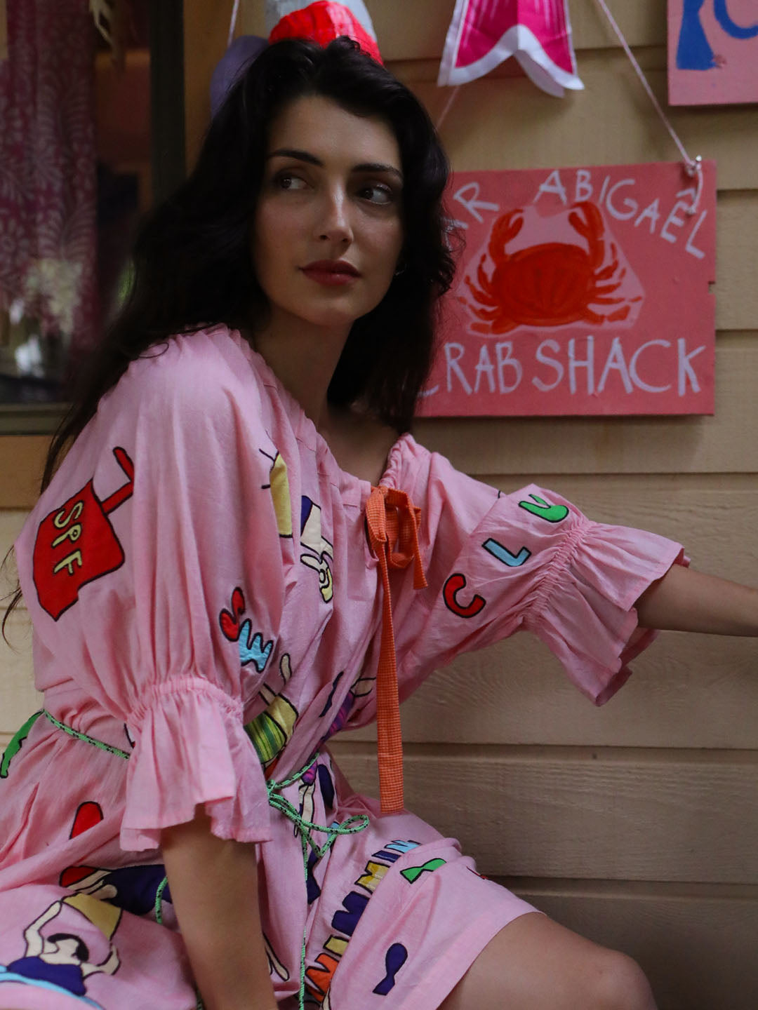 A woman wearing a pink short-sleeve beach cover-up with colorful appliqué swimming figures and an orange tie, seated in front of a wooden wall with a 'Crab Shack' sign.