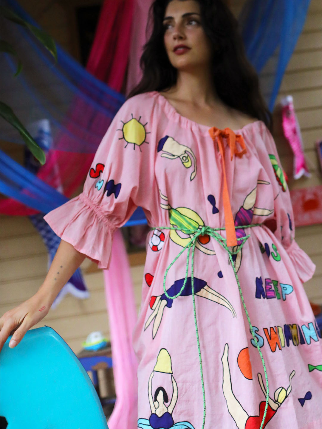 A woman wearing a light pink off-the-shoulder dress with colorful appliqué swim and sun motifs, standing in a decorated indoor setting with draped pink and blue fabric.