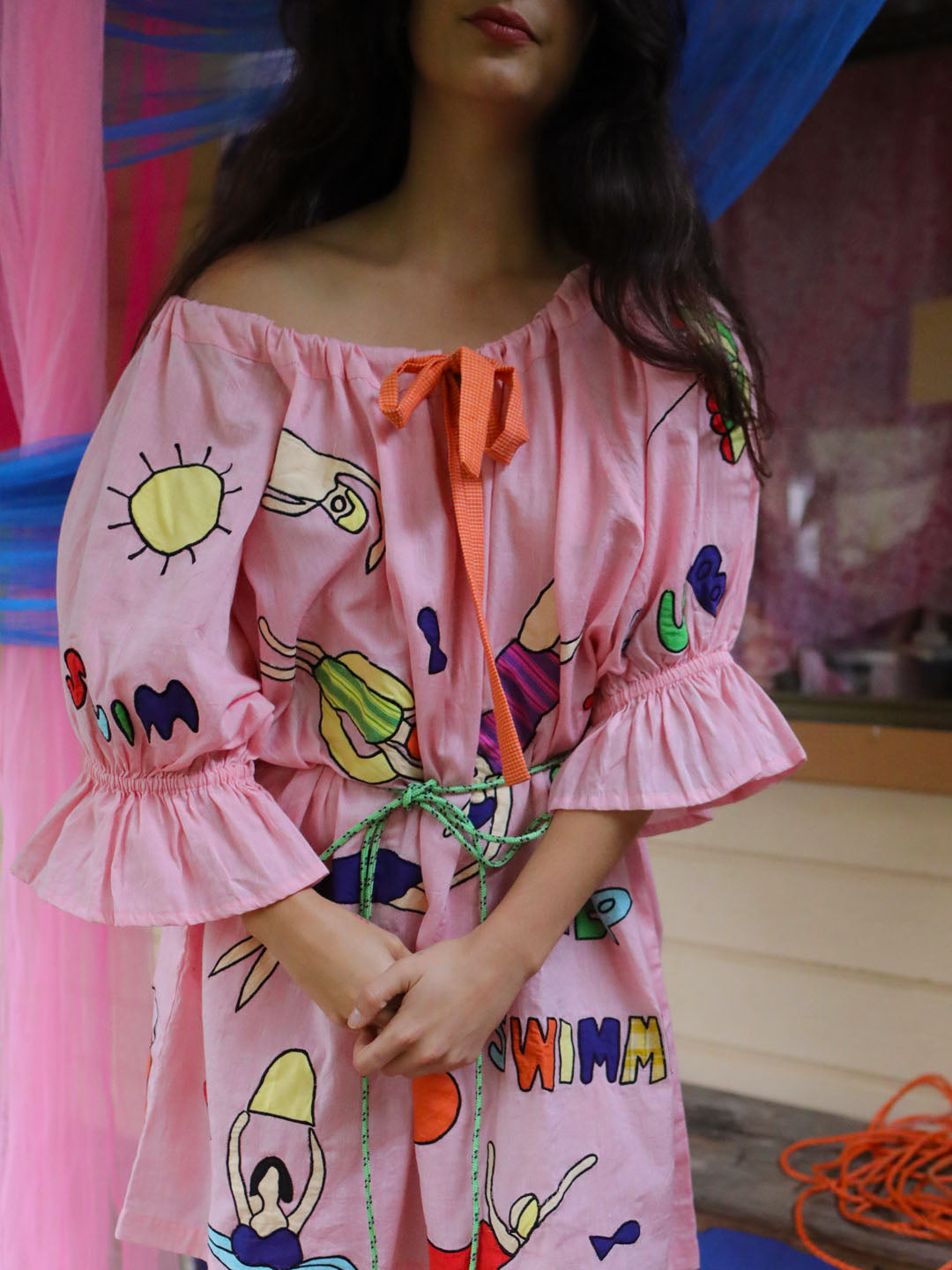 A woman wearing a pink off-the-shoulder dress with colorful appliqué swimming figures and an orange tie at the neckline, photographed indoors against a soft pink and blue draped background.