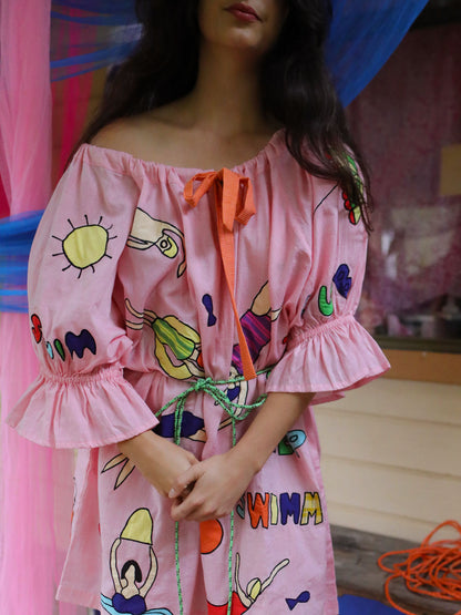 A woman wearing a pink off-the-shoulder dress with colorful appliqué swimming figures and an orange tie at the neckline, photographed indoors against a soft pink and blue draped background.