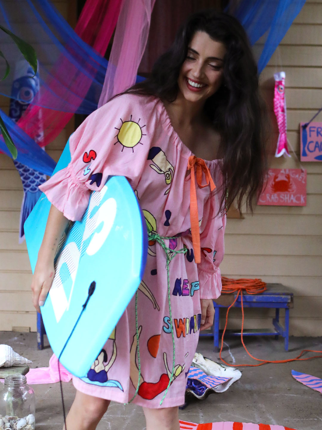 A woman wearing a pink off-the-shoulder swimsuit dress with colorful appliqué swimming figures and holding a light blue surfboard, standing in a decorated outdoor patio setting.