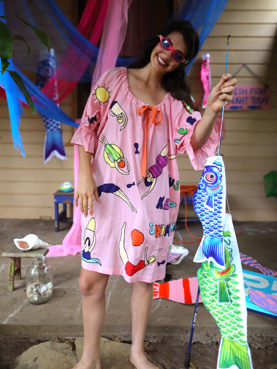 A woman wearing pink swimming goggles, a pink short-sleeve kaftan-style dress with colorful swim and sun illustrations, standing barefoot on a porch while holding a large blue koi fish decoration and fishing rod.