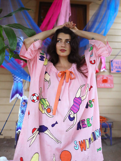 A woman wearing a pink kaftan-style cover-up dress with colorful swim-themed illustrated appliqué and text, standing outdoors in front of a house with colorful fabric decorations.