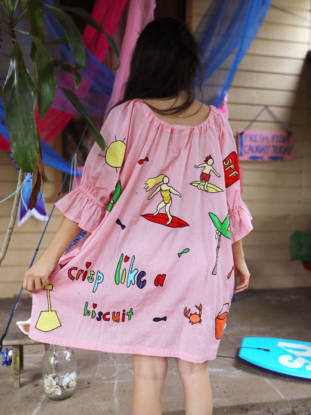 A woman viewed from behind wearing a pink short-sleeve beach cover-up dress with colorful beach-themed appliqué prints and the phrase 'crisp like a biscuit' on the back, standing outdoors near a wooden wall and hanging fabric.