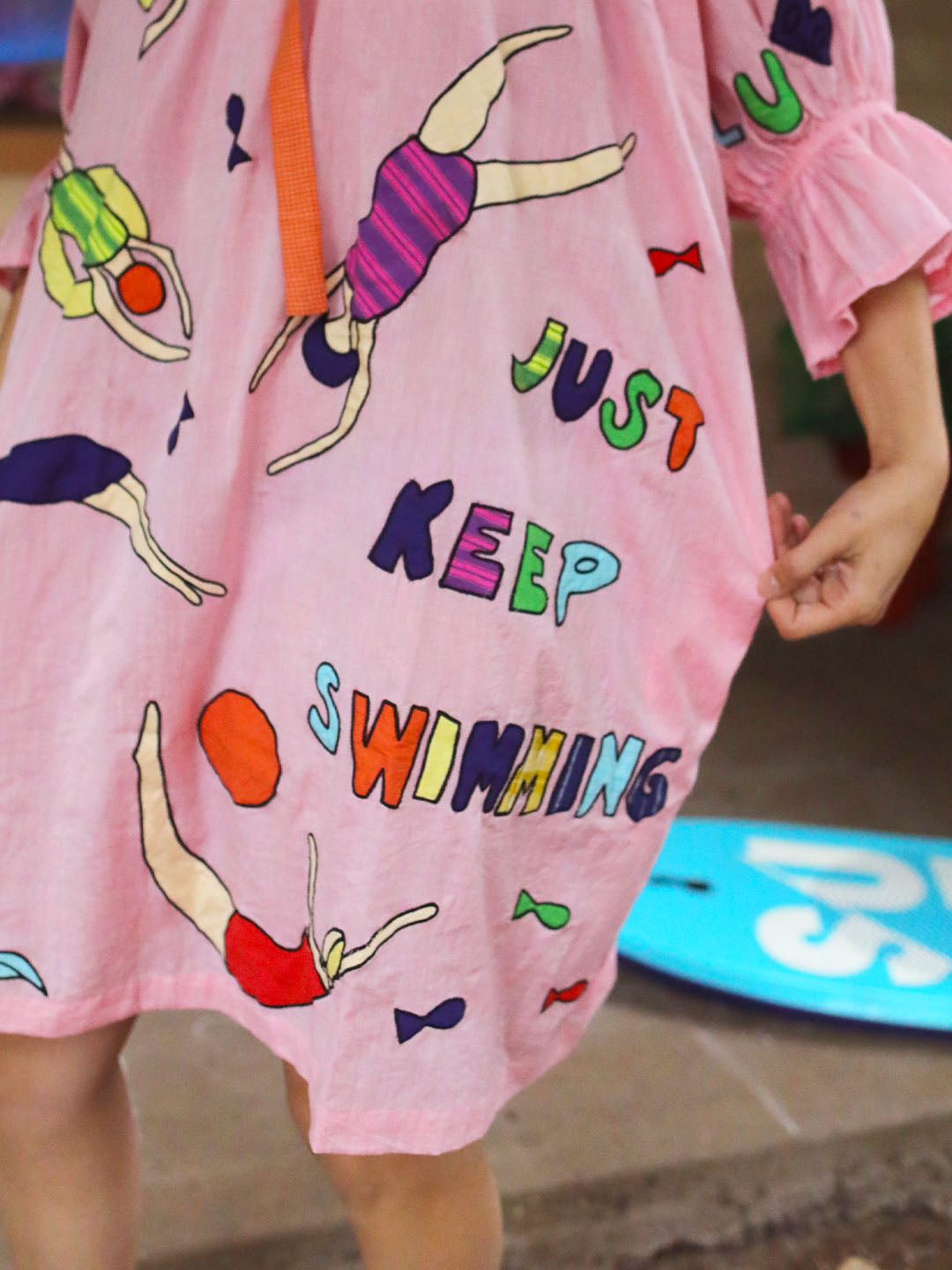 A close up of pink short-sleeve dress with a colorful 'Just Keep Swimming' graphic print, shown on a woman in an outdoor setting.