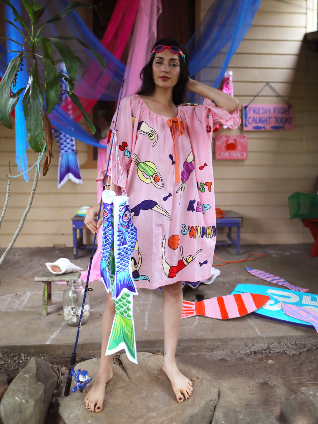 A woman stands barefoot outdoors wearing a loose pink beach cover-up  dress decorated with colorful appliqué swimming figures. She holds a large fish-shaped decoration on blue fishing rod, and other decorative items and fabric hangings are visible in the background.