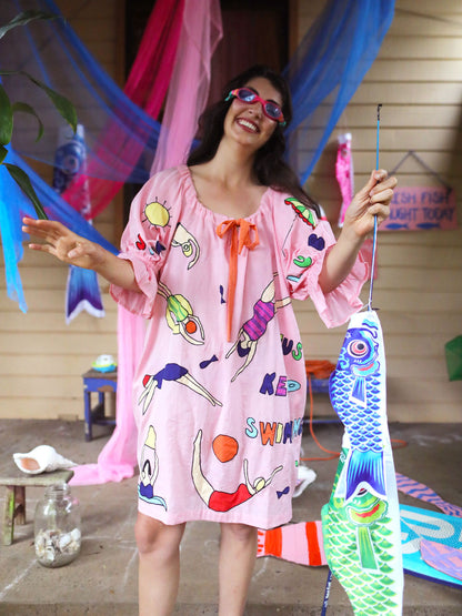 A woman wearing pink swimming goggles, a pink swim-themed smock dress with colorful hand drawn appliqué swimmer print and holding a large koi fish decoration, standing on a porch decorated with pink and blue fabric.