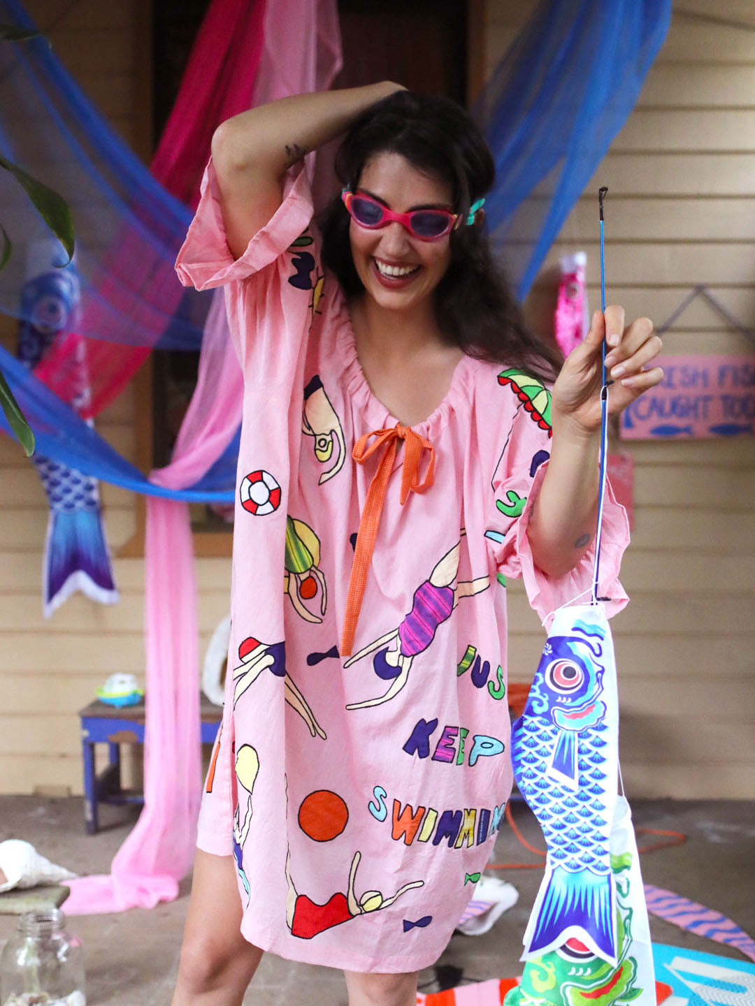A woman wearing a pink swim-themed smock dress with colorful hand drawn appliqué swimmers and an orange bow, holding a decorative fish-shaped flag. She is posing in front of a house with pink and blue fabric decorations.