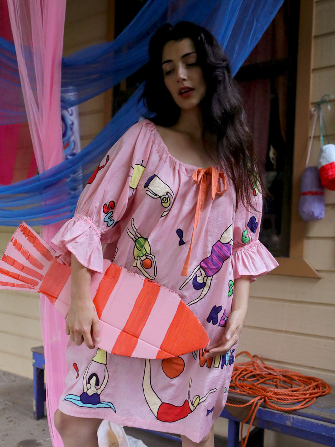 A woman wearing a pink off-the-shoulder dress with a colorful appliqué swimming figures print, holding a pink and orange striped painted fish prop in front of a porch with blue and pink drapes.