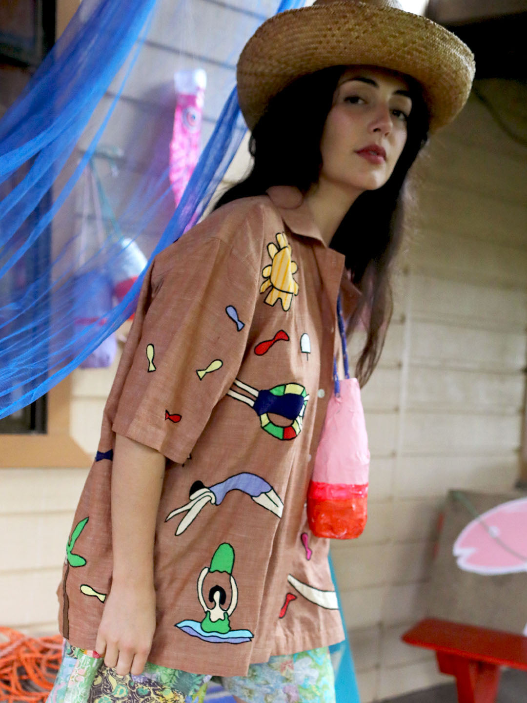 A woman wearing a straw hat and a brown short-sleeve button-up shirt with colorful retro-style cartoon graphics, standing in a decorated outdoor setting with a blue net and a pink bag.