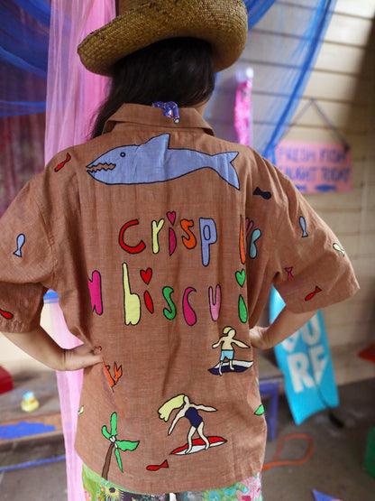 A person viewed from behind wearing a tan short-sleeve button-up shirt with colorful hand-drawn graphics, including a blue shark, the words 'Crisp like a Biscuit,' and surfers, along with a straw hat.