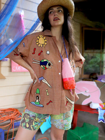 A woman wearing a brown short-sleeve button-up shirt with colorful beach-themed illustrations, a straw hat, and patterned shorts, standing in a casual outdoor setting.