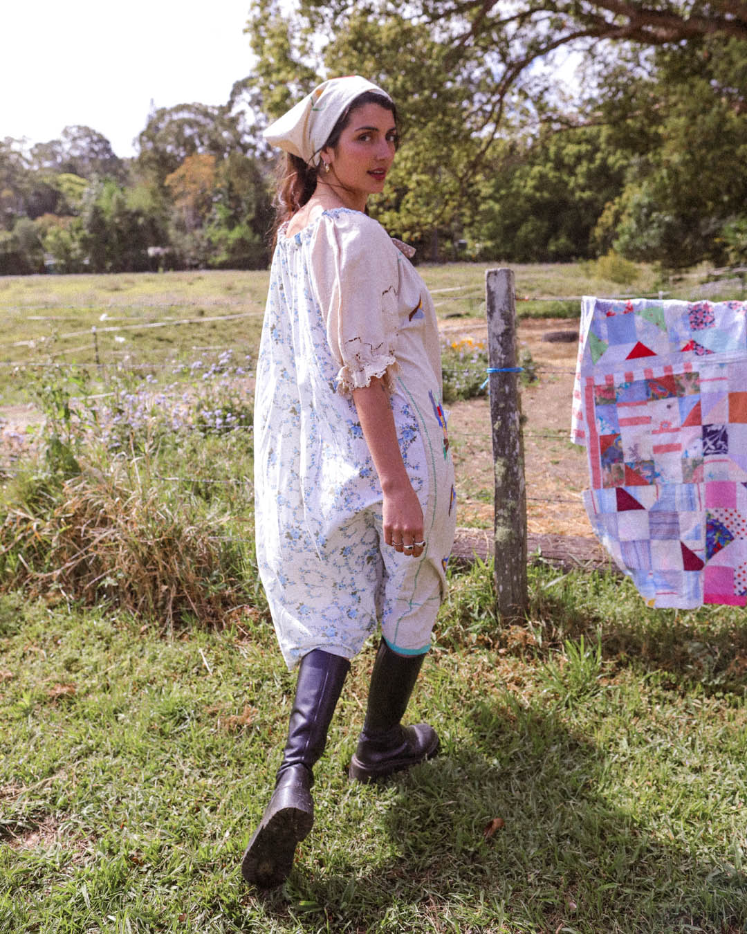 A woman wearing a light beige short-sleeved dress with a blue floral print, a matching headscarf, and black rubber boots stands in a grassy outdoor field next to a patchwork quilt hanging on a fence post.