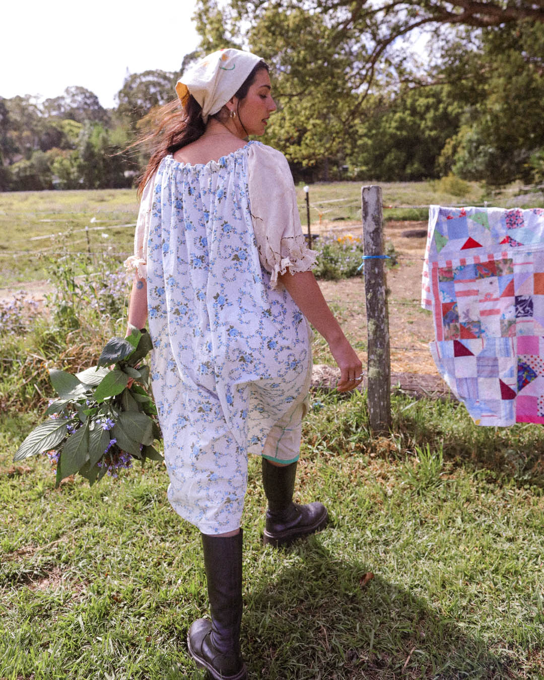 A woman stands outdoors in a grassy field wearing a loose white dress with a blue floral print, a cream puff-sleeve blouse, and dark rubber boots. A colorful patchwork quilt hangs on a wooden post beside her.