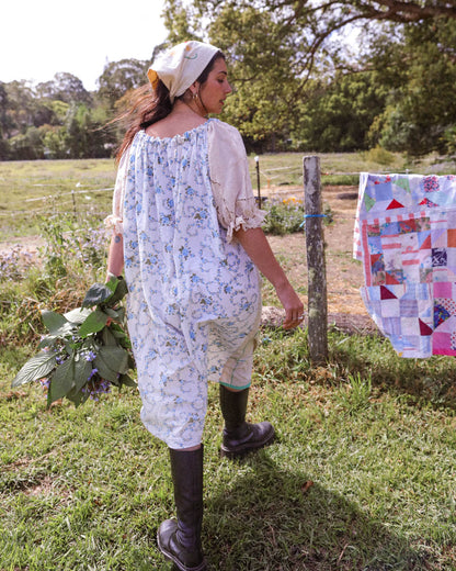 A woman stands outdoors in a grassy field wearing a loose white dress with a blue floral print, a cream puff-sleeve blouse, and dark rubber boots. A colorful patchwork quilt hangs on a wooden post beside her.