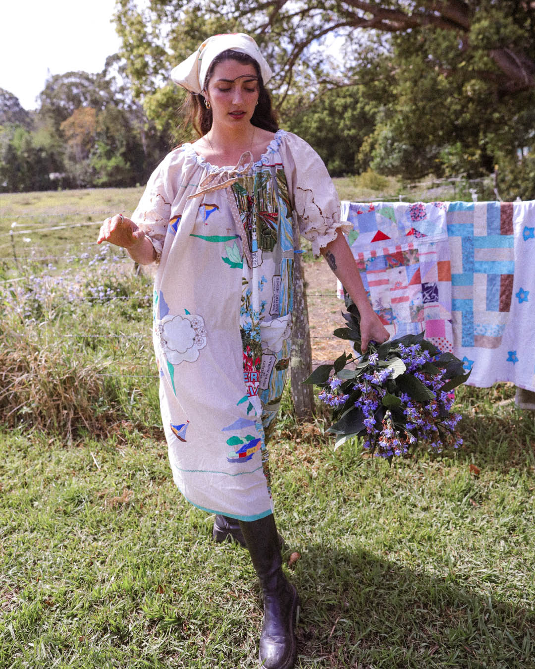 A woman stands outdoors in a grassy field wearing a loose, cream-colored dress with colorful abstract and floral patterns, holding a bouquet of purple flowers. A quilt is hanging on a tree in the background.