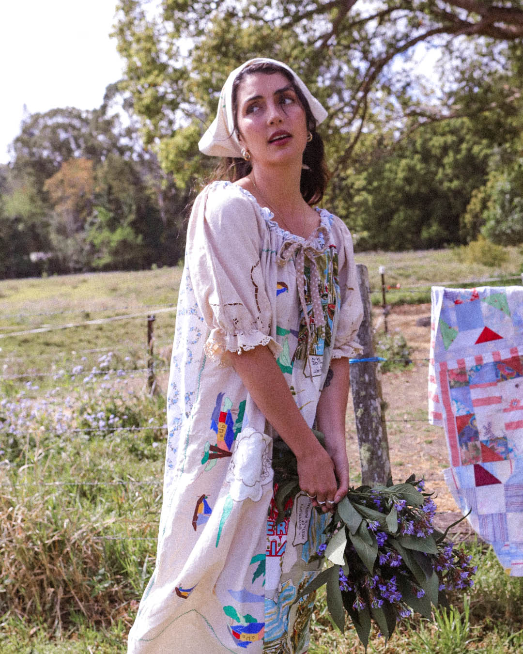 A woman wearing a vintage-style cream dress with colorful printed motifs and a matching bonnet stands outdoors in a grassy field, holding a bouquet of purple flowers. A quilted textile hangs on a fence post to her right.