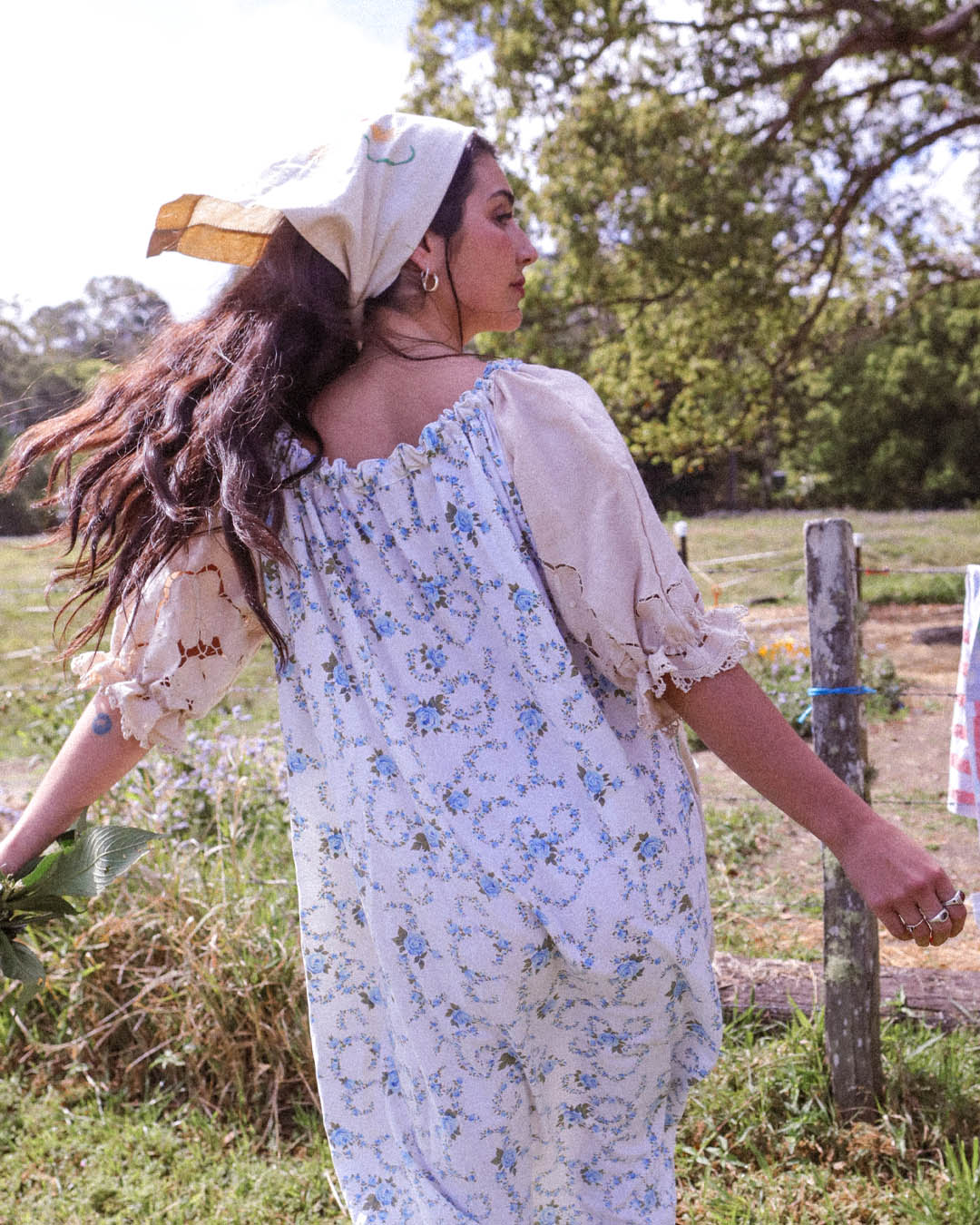 A woman wearing a loose, short-sleeved floral dress with a cream headscarf, photographed outdoors in a grassy field with trees in the background.