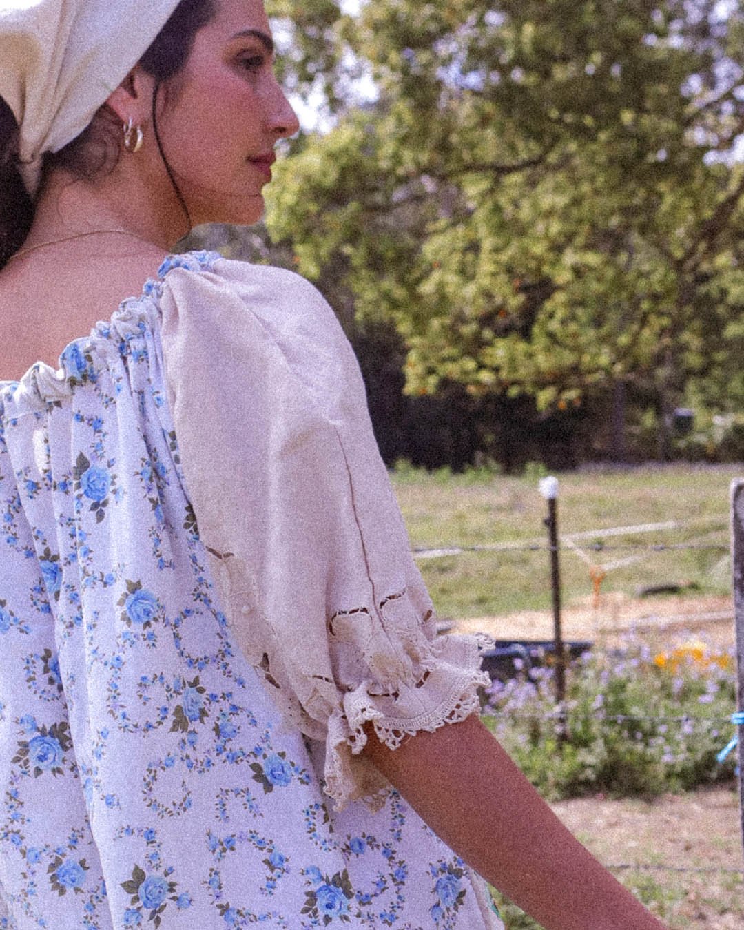 A woman wearing a light beige headscarf and a white puff-sleeve blouse with a blue floral print, photographed outdoors in a sunny, grassy field.