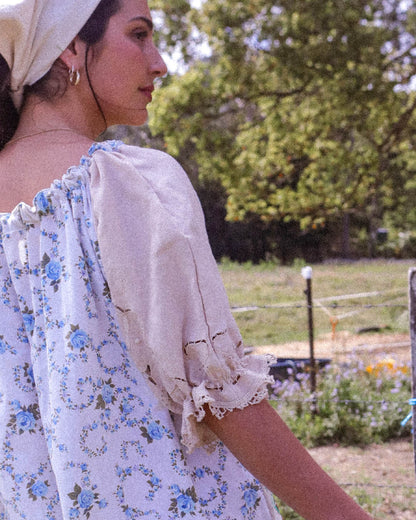 A woman wearing a light beige headscarf and a white puff-sleeve blouse with a blue floral print, photographed outdoors in a sunny, grassy field.