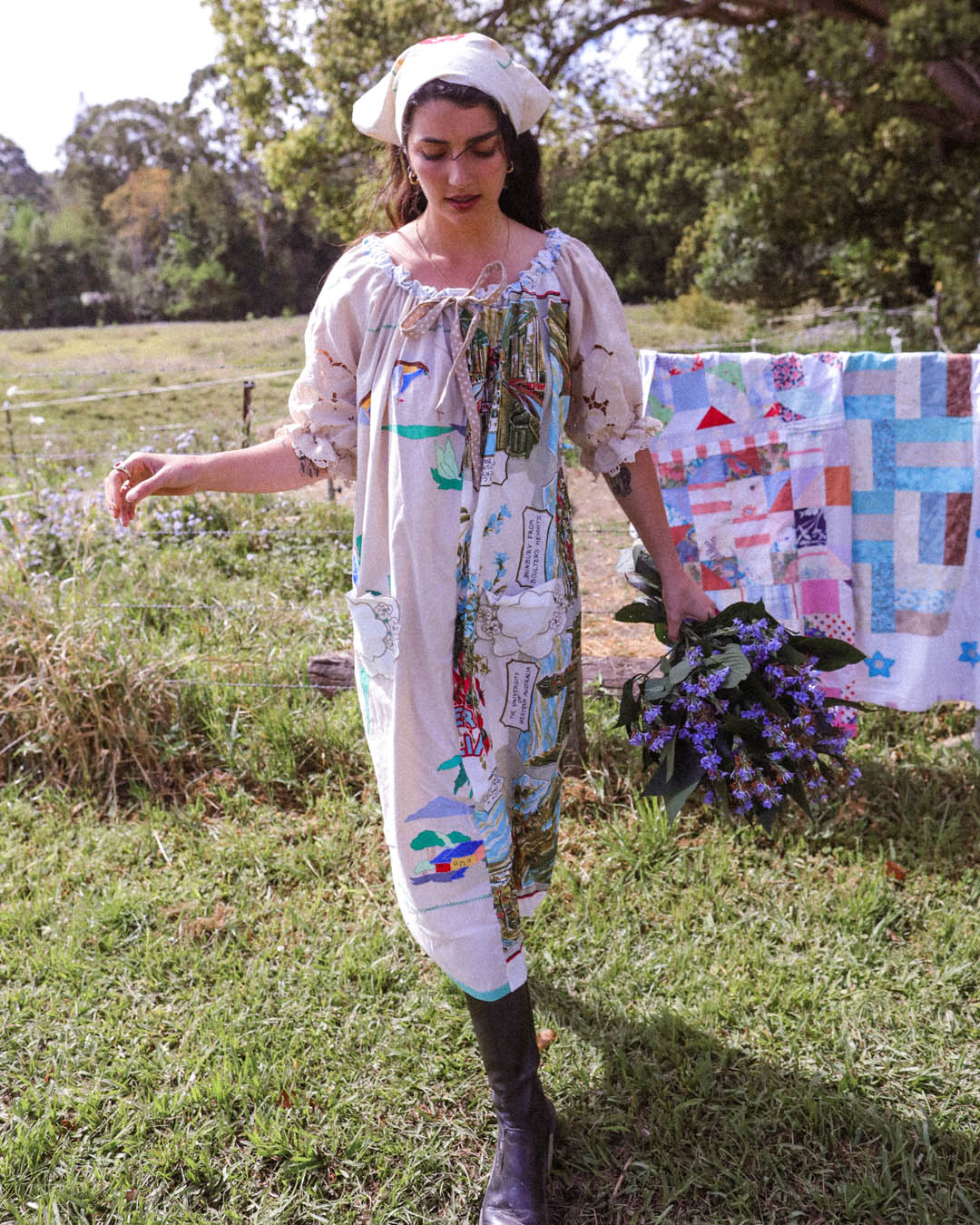 A woman wearing a long-sleeved, knee-length patchwork dress with a white hat and dark boots stands in a grassy outdoor field. She is holding a bouquet of purple flowers, with a colorful patchwork quilt hanging in the background.