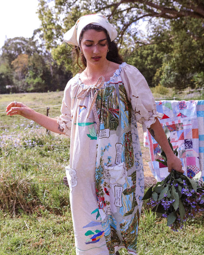 A woman standing outdoors in a grassy field wearing a long, loose white dress with colorful botanical prints and tie details, holding a bouquet of purple flowers. A quilt is visible on a clothesline in the background.