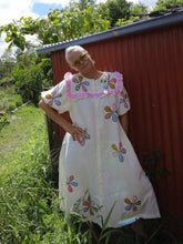 Load image into Gallery viewer, Woman wearing a white dress with colorful floral embroidery standing in front of a red building.